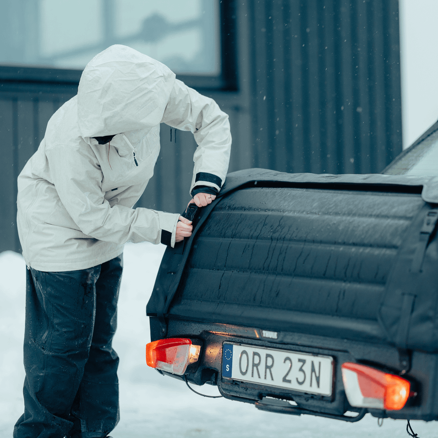 A person wearing a hooded winter coat secures the Thule Onto 2 cargo bag onto the towbar of a car ensuring a firm fit in a cold, snowy setting.